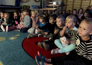 Dzieci siedzą na dywanie i jedzą popcorn.