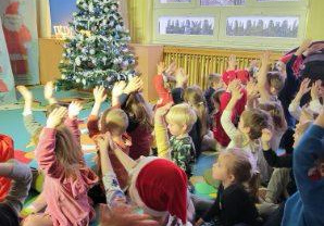 Przedstawienie świąteczne. Dzieci siedzą na dywanie i klaszczą w ręce nad głową. Z tyłu widać choinkę ubraną w białe ozdoby.