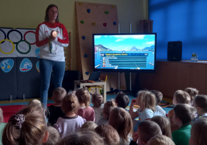 Dzieci słuchają zawodniczki olimpijskiej i oglądają na ekranie wyścig kajakarski.