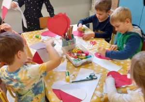 Dzieci siedzą przy stoliku i wykonują chorągiewki z papieru w kształcie serca.