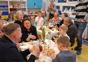 Dzień Babci i Dziadka. Dziadkowie, babcie i przedszkolaki siedzą przy stolikach i jedzą słodkości.