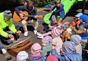 Koncert. Dzieci siedzą na placu zabaw i słuchają, jak pan gra łyżeczkami na szprychach koła.