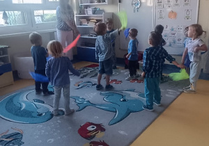 Dzieci wymachują kolorowymi chustami stojąc na dywanie.