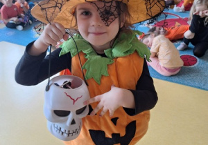 Dziewczynka w stroju dyni, z kapeluszem czarownicy na głowie i z koszykiem w kształcie czaszki.