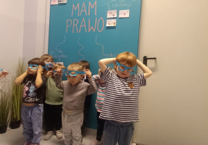Dzieci stoją pod ścianą i przymierzają niebieskie okulary.